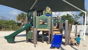 Eine Spielkonstruktion für Kinder mit Rutschen und Kletterelementen, auf sandigem Untergrund mit Palmen im Hintergrund.