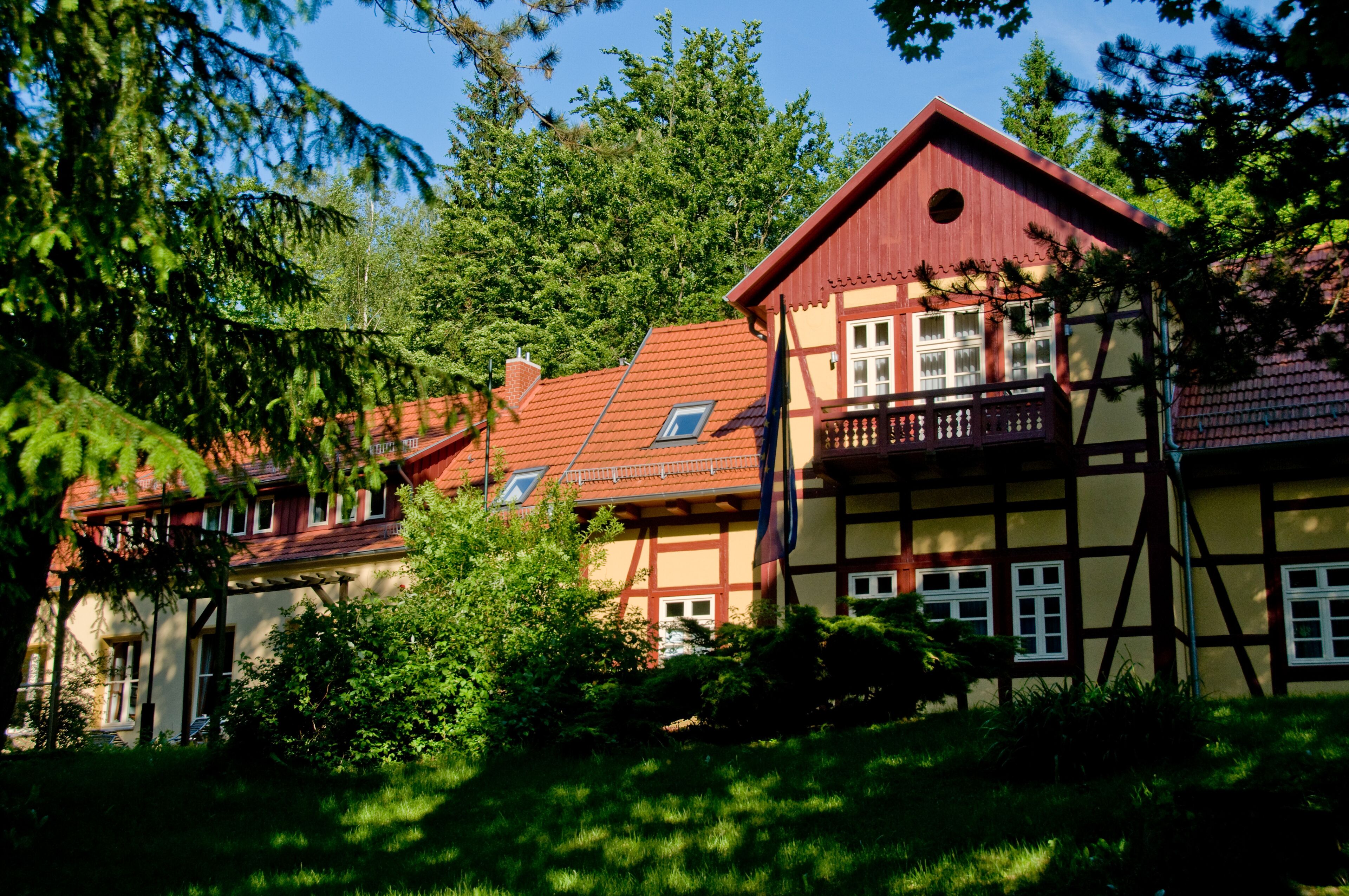 Une maison traditionnelle à colombages, avec un toit rouge, entourée d’une végétation luxuriante et d’arbres.