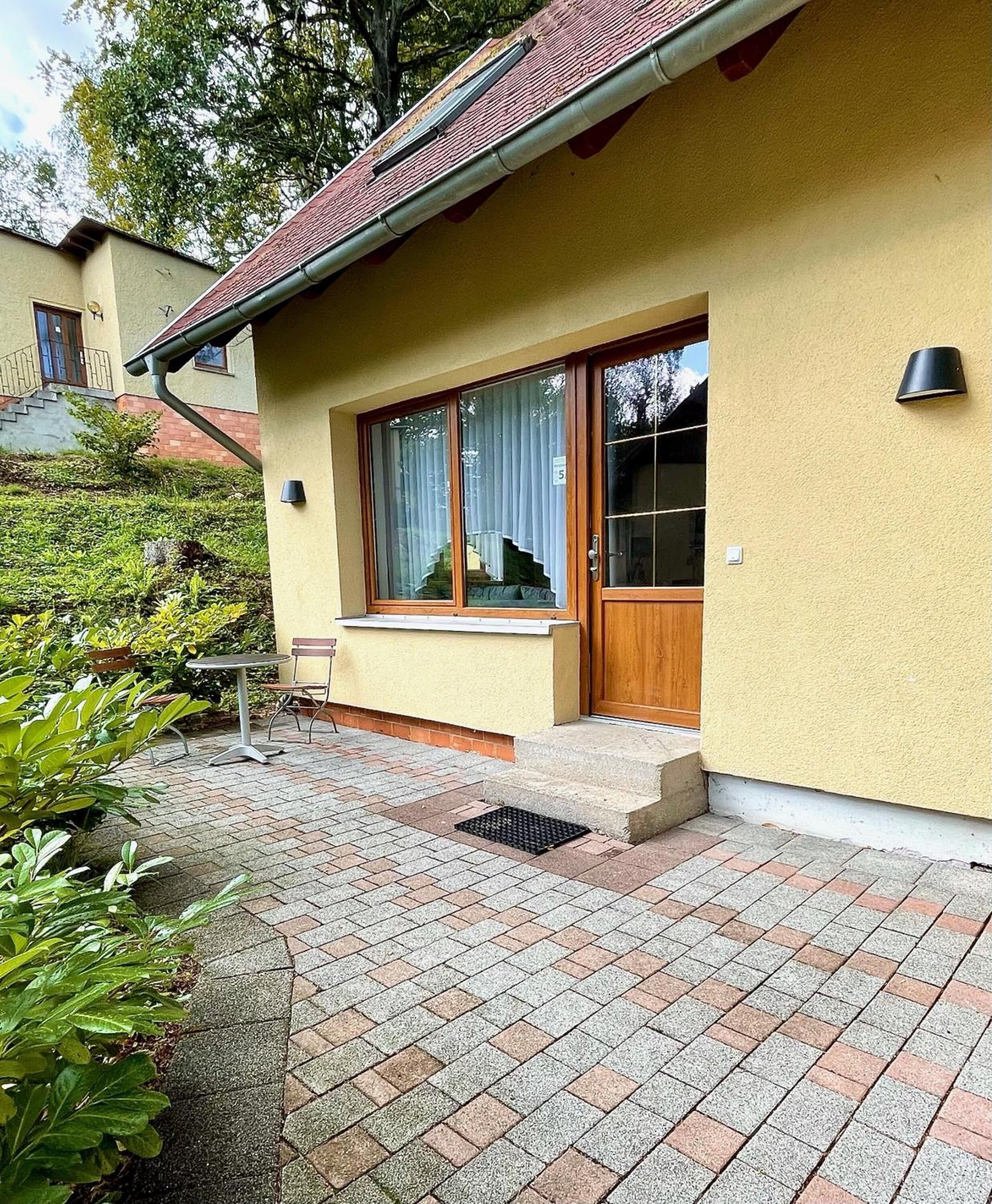 A house with a yellow exterior, a wooden door, and a window with a curtain. There is a small outdoor seating area with a table and chairs.