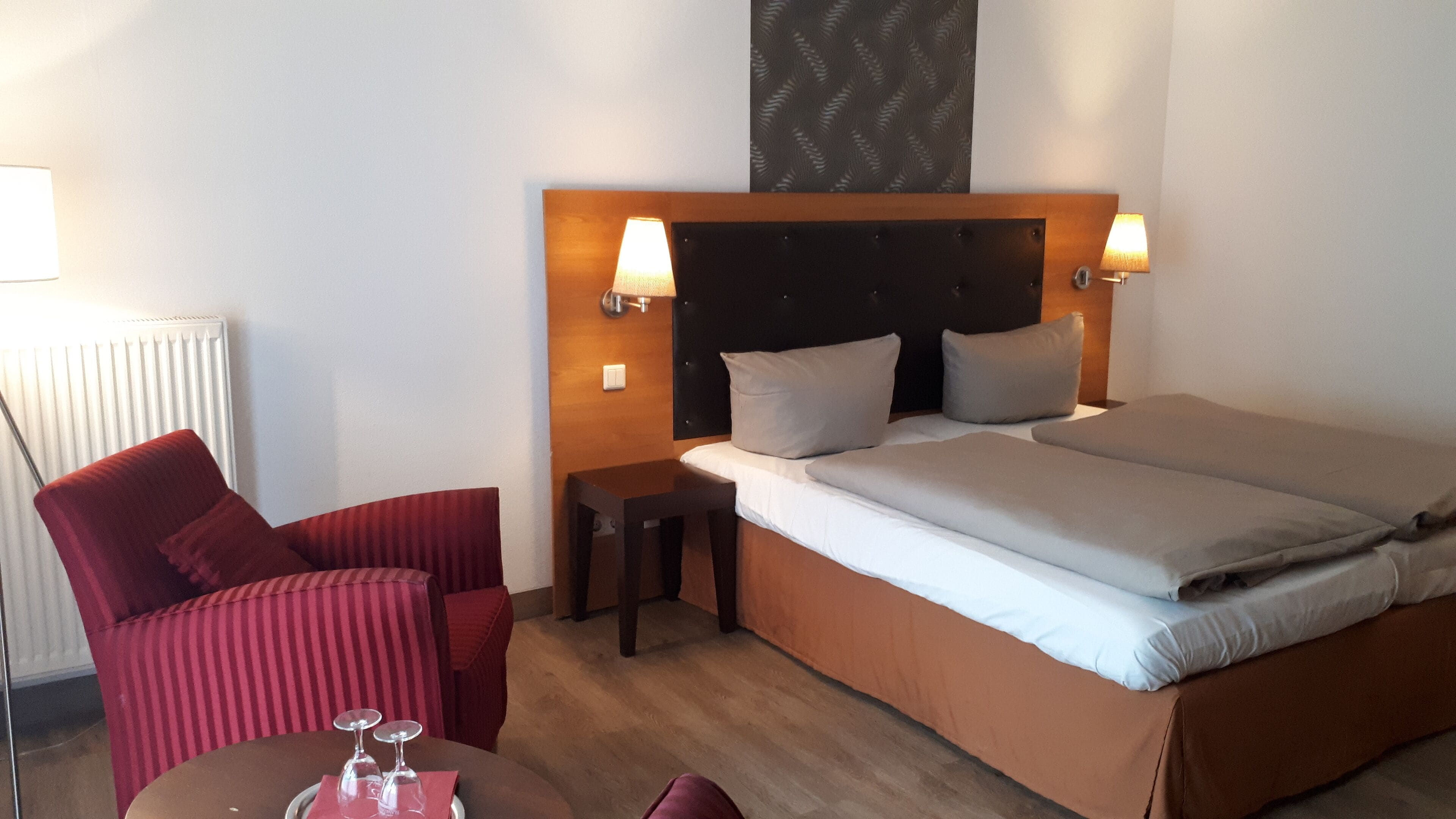 Una camera d'albergo con un letto, due lampade da comodino, una poltrona rossa, un tavolino con dei bicchieri e un termosifone.