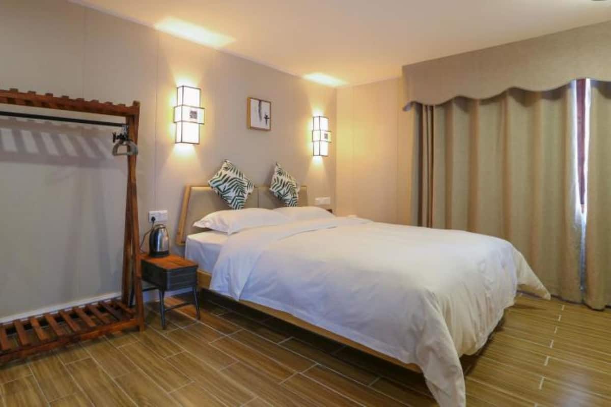 Habitación de hotel con una cama, un armario de madera, una mesita de noche, dos lámparas de pared y una ventana con cortinas.