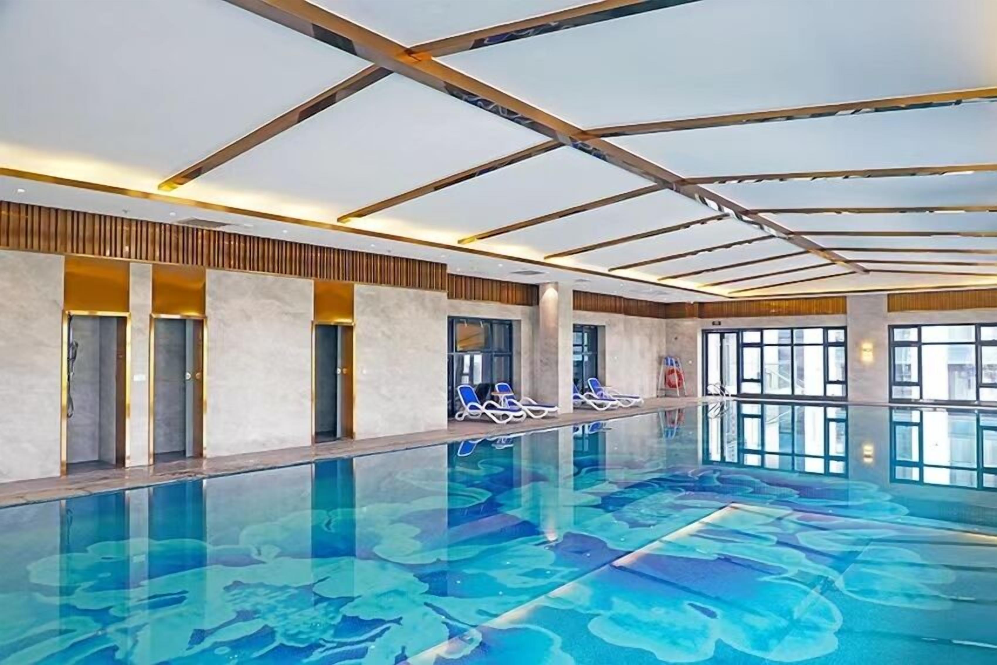 An indoor swimming pool with clear water, lounge chairs, and a modern ceiling design.