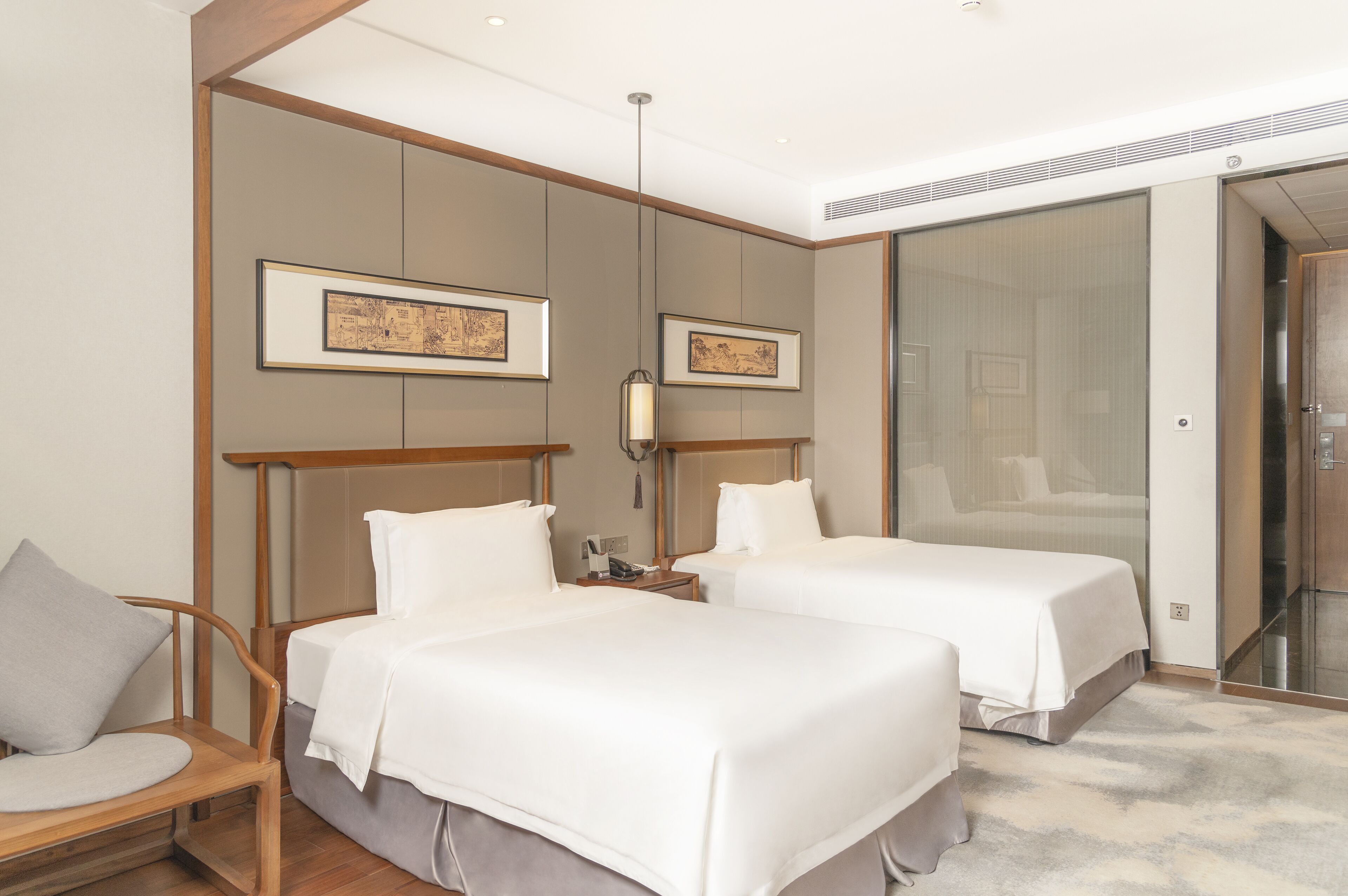 Una habitación de hotel con dos camas, una silla de madera, una mesita de noche y cuadros enmarcados en las paredes.
