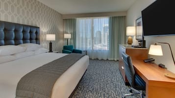 Quarto de hotel com uma cama grande, uma escrivaninha com cadeira, TV de tela plana e uma janela com cortinas.