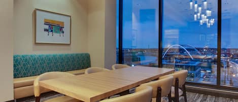 A modern hotel room with a large window offering a city view, a wooden dining table, and a green patterned bench.