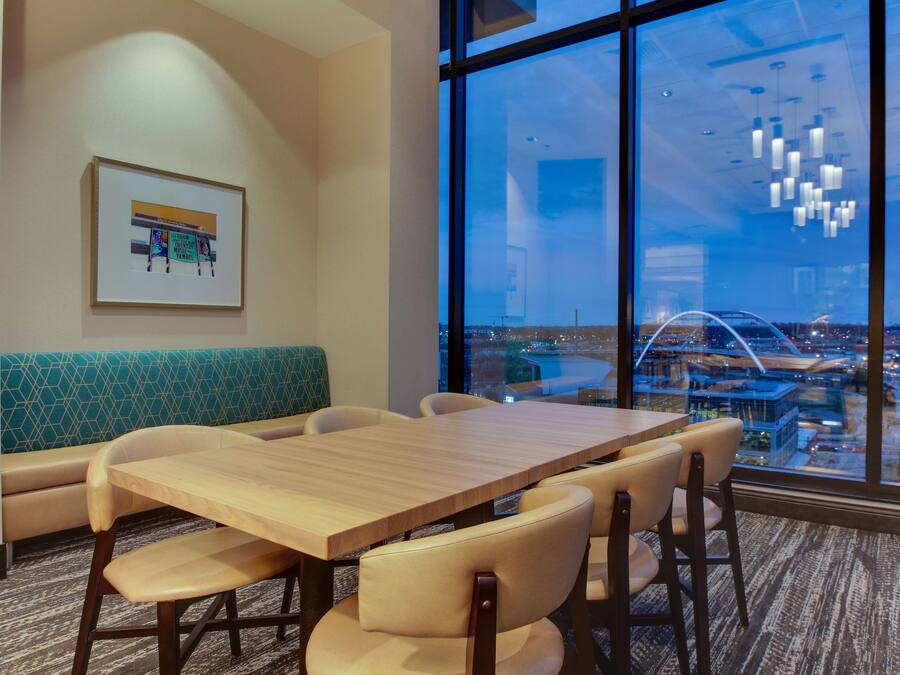 A modern hotel room with a large window offering a city view, a wooden dining table, and a green patterned bench.