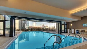 A modern indoor swimming pool with clear glass walls, a tiled floor, and a view of a cityscape with buildings and cranes. - Drury Plaza Hotel Nashville Downtown (Nashville)