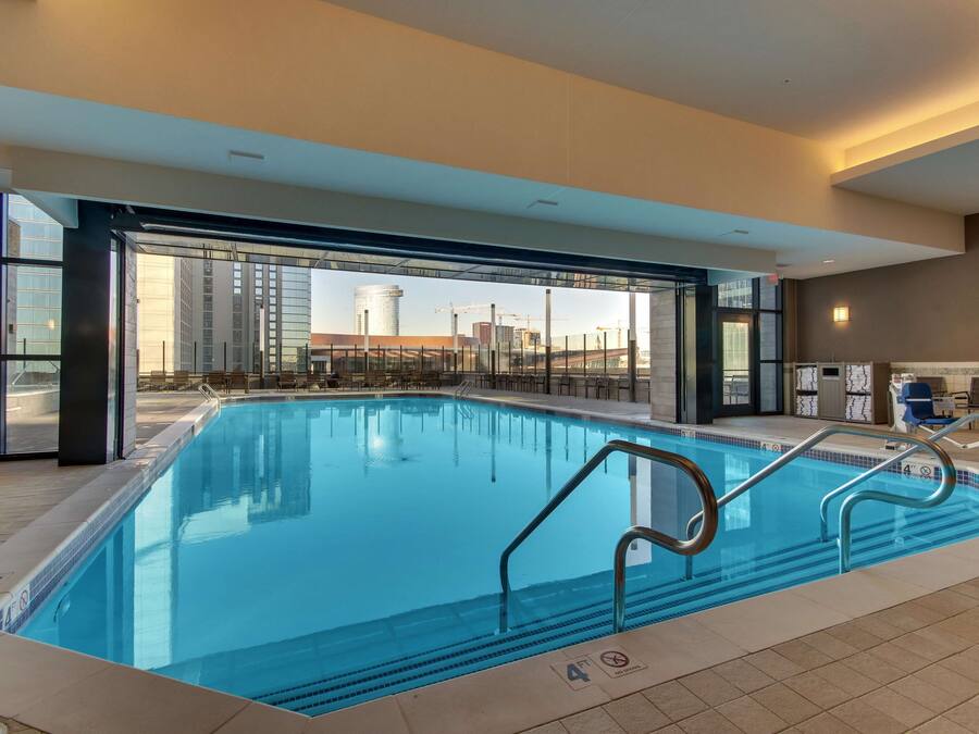 Piscina coberta moderna com paredes de vidro transparente, chão revestido e vista para um horizonte urbano com edifícios e guindastes.