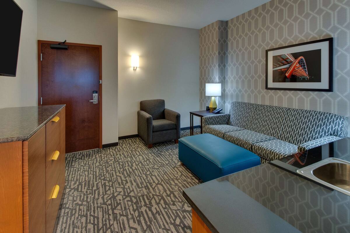 Une chambre d’hôtel avec un bureau en bois, un fauteuil gris, un canapé à motifs, un pouf bleu, une petite table avec une lampe et un tableau encadré au mur.