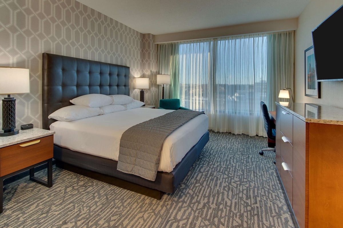 Une chambre d’hôtel avec un grand lit, deux tables de chevet avec des lampes, un bureau avec une chaise, un téléviseur fixé au mur et une fenêtre avec des rideaux.