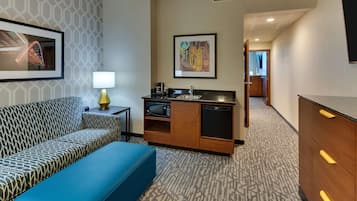 Ein Hotelzimmer mit einer gemusterten Couch, einem blauen Ottomanen, einer Küchenzeile mit Mikrowelle und Kaffeemaschine sowie gerahmten Kunstwerken an den Wänden.