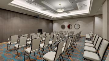 Sala de conferência com fileiras de cadeiras, um projetor e teto com iluminação embutida e claraboia.