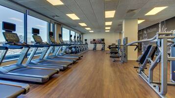 Uma academia com esteiras alinhadas junto às janelas, piso de madeira e equipamentos de exercícios à direita.