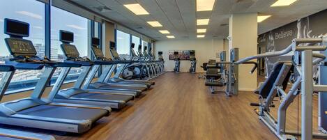 A gym with treadmills aligned along the windows, a wooden floor, and exercise equipment on the right.
