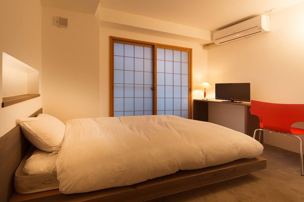 Een slaapkamer met een bed, een bureau met stoel, een televisie en een airconditioning.