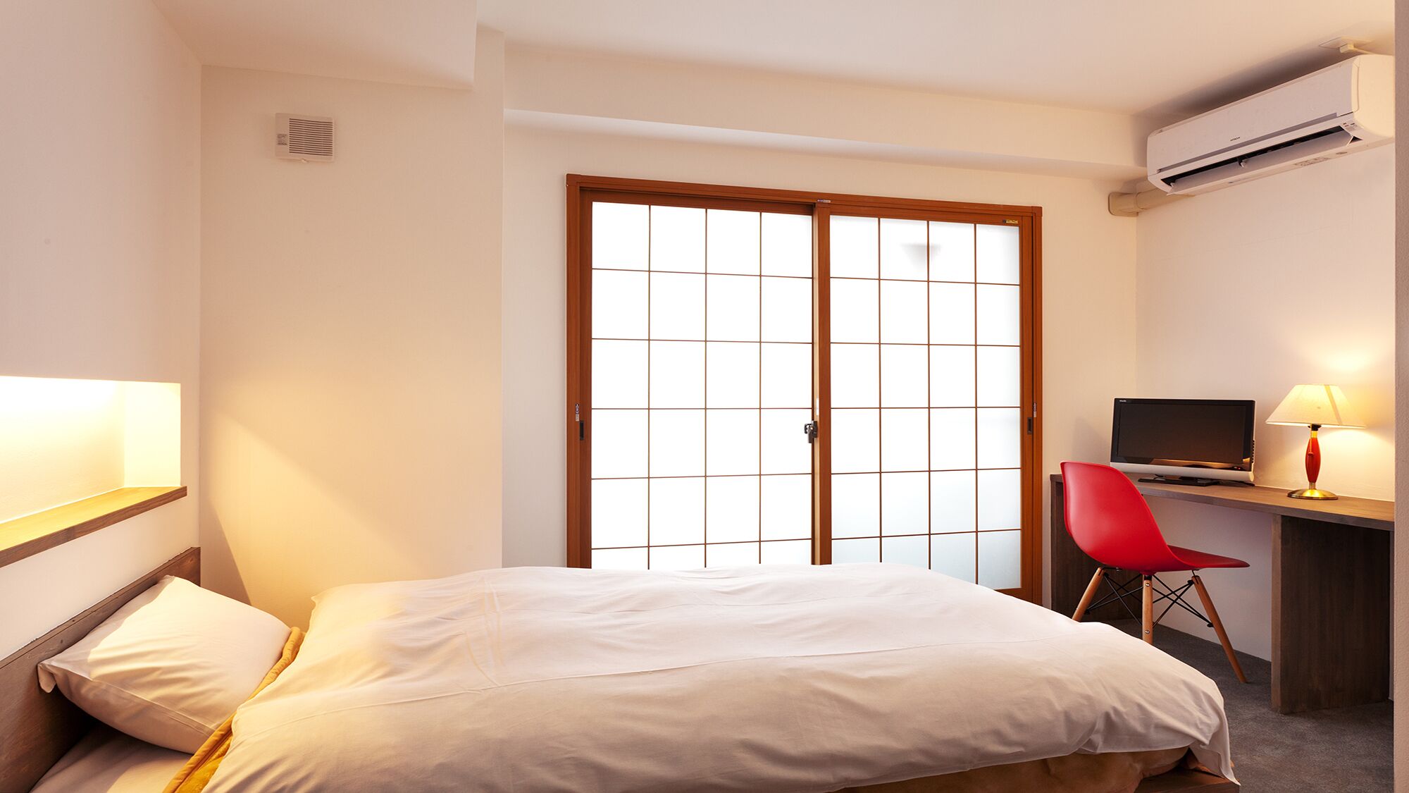 Una habitación de hotel con una cama, un escritorio con computadora, una silla roja y un equipo de aire acondicionado.