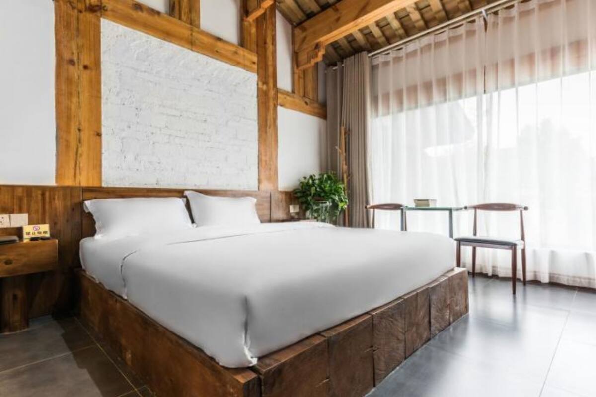 Ein Schlafzimmer mit einem großen Bett, Holzbalken, einer weißen Ziegelwand, einer Pflanze und einem Fenster mit Vorhängen.