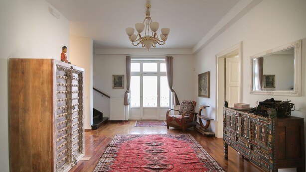 A spacious room with a wooden floor, a large red patterned rug, a chandelier, a wooden cabinet, and a chair with a patterned cushion. - Hotel Vila Katrca (Ljubljana)