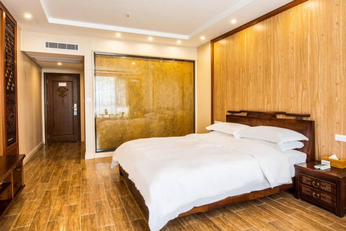 Una habitación de hotel con piso de madera, una cama grande con ropa blanca, un cabecero de madera y una mesita de noche con lámpara.