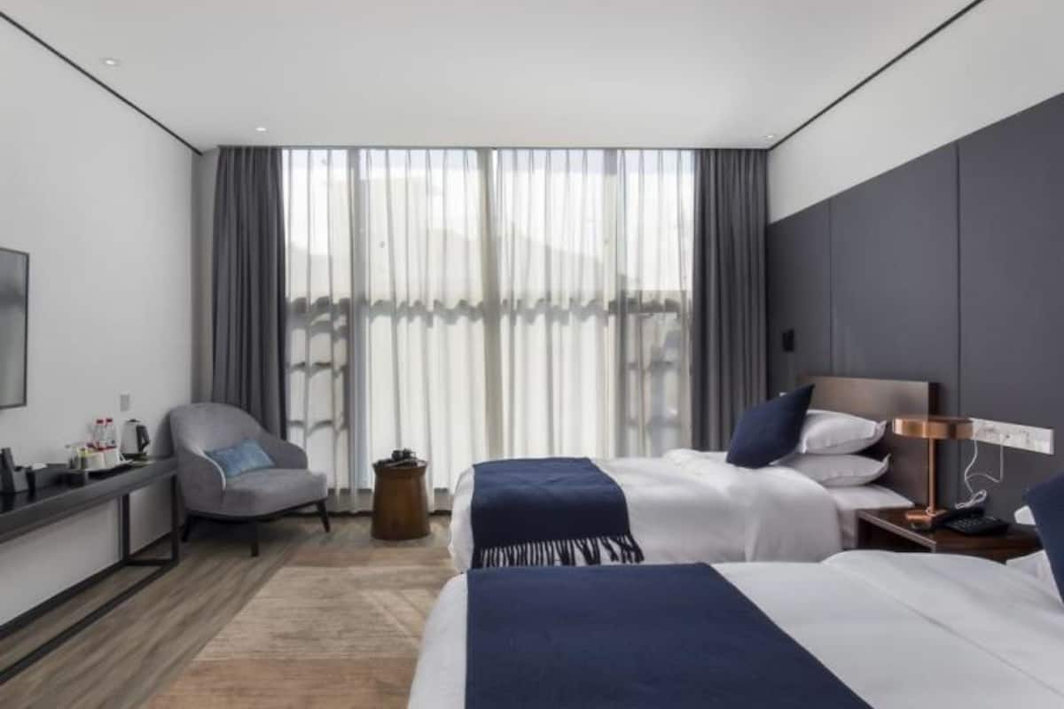 A modern hotel room with two beds, a desk, a chair, and a large window with curtains.