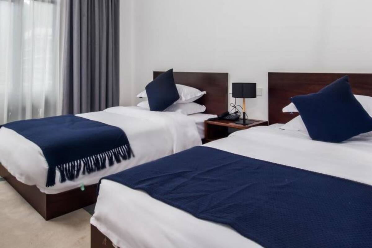 A hotel room with two beds, each with a dark blue bedspread and white linens. There is a nightstand with a lamp and a phone on one side.