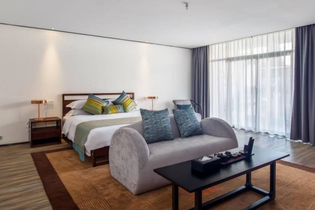 A modern hotel room with a bed, a sofa, a coffee table, and a large window with curtains.