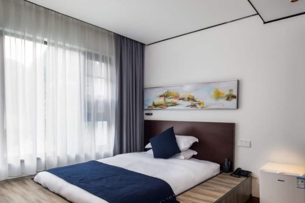A modern hotel room with a wooden floor, a bed with white and navy bedding, a dark headboard, and a painting above the bed.