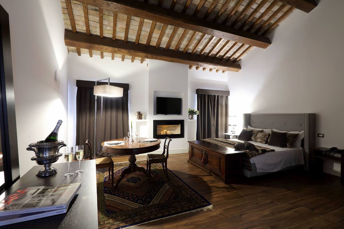 A room with a wooden ceiling, a bed with a white headboard, a dining table with a rug, and a fireplace.
