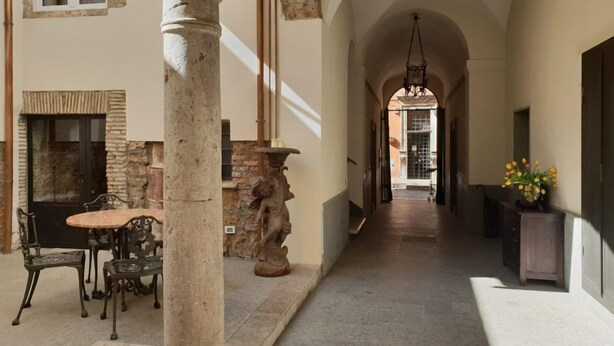 Un pasillo con una columna de piedra, un banco, una mesa con sillas y una estatua.