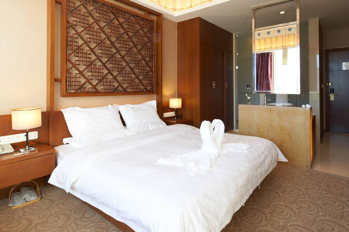 A hotel room with a large bed, two bedside tables with lamps, a wooden headboard, a decorative wall panel, a bathroom with a mirror and sink, and a door.