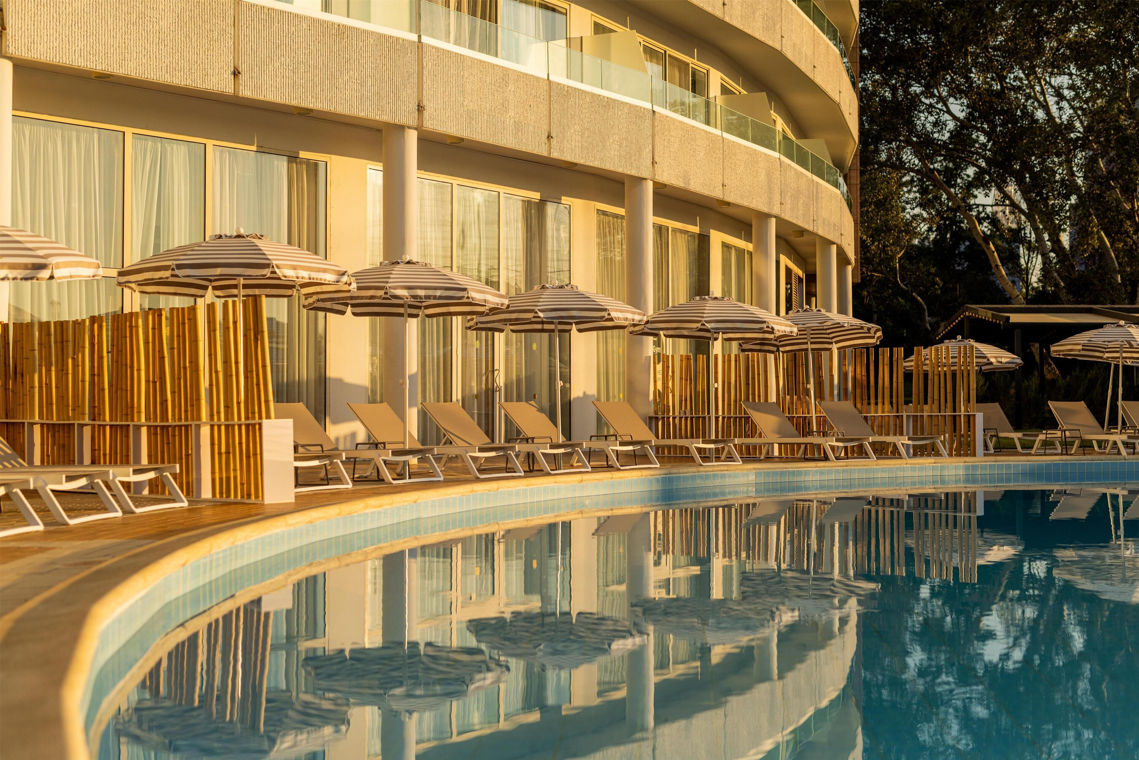 A hotel pool area with sun loungers and umbrellas, surrounded by a building with large windows and a reflection in the pool.