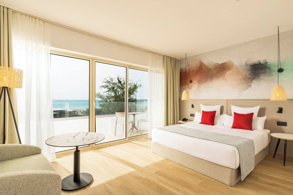A hotel room with a bed, a round table, a sofa, a large window with a view, and a painting on the wall.