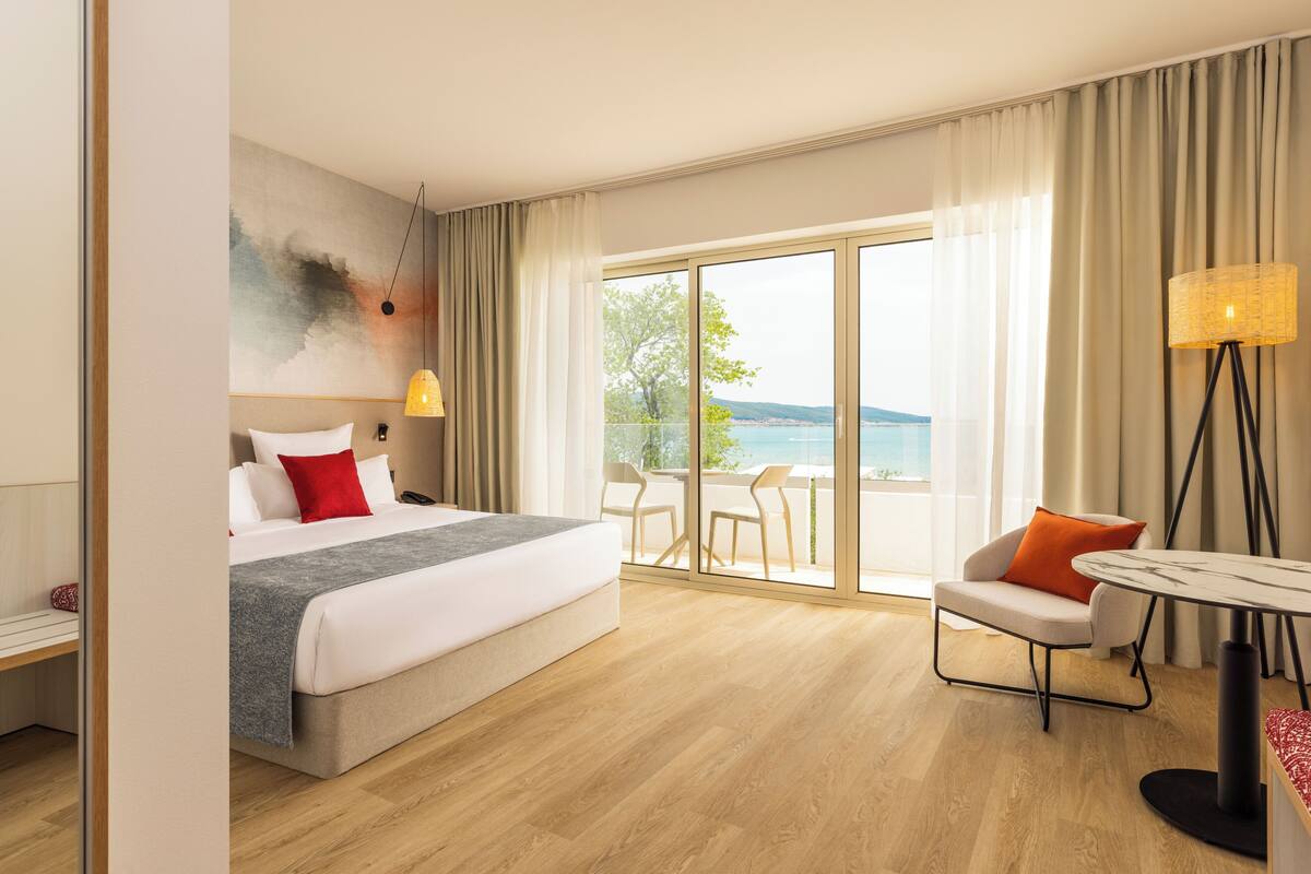 A hotel room with a bed, a chair, a table, and a view of the sea.