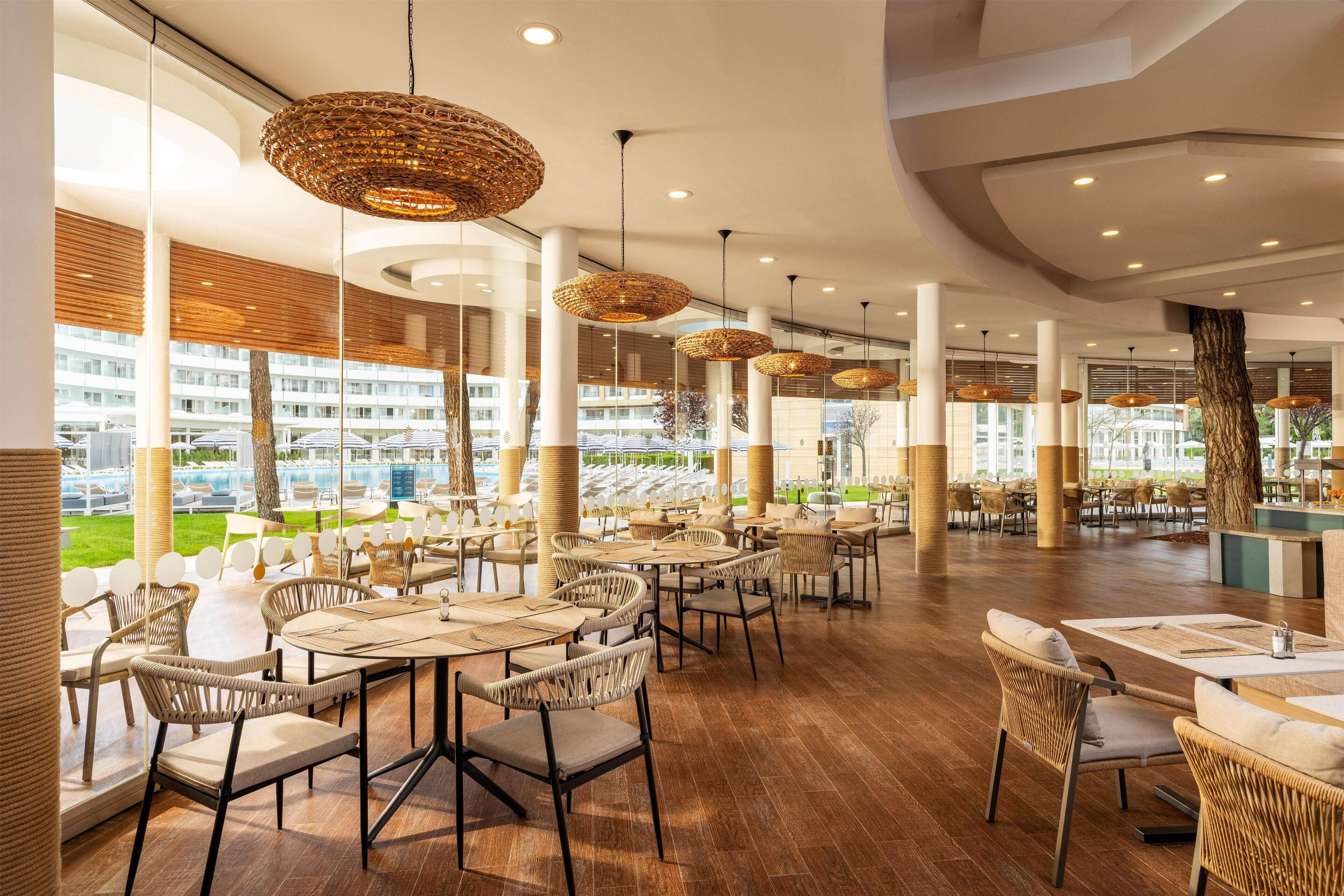 A spacious indoor dining area with wicker chairs and tables, large windows offering views of a building and greenery outside.