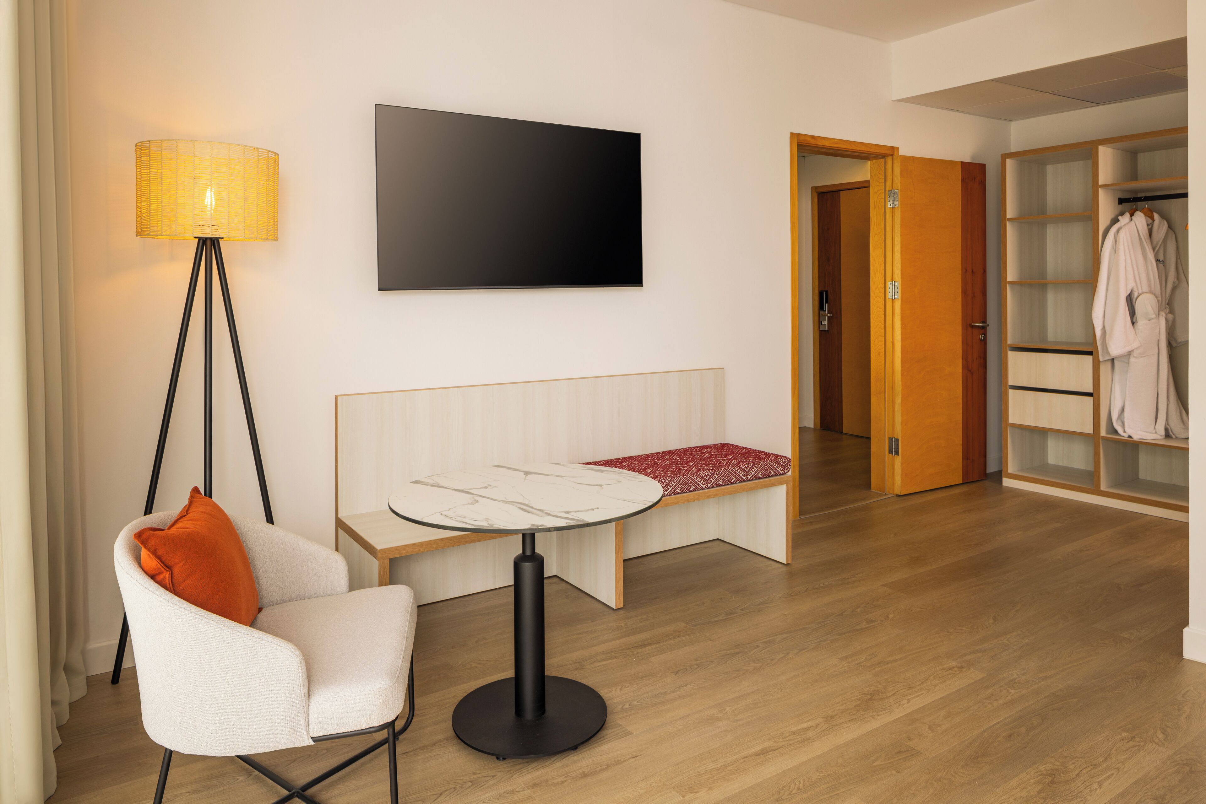 A modern hotel room with a bed, a bedside table, a chair, a floor lamp, a TV, and a wardrobe.