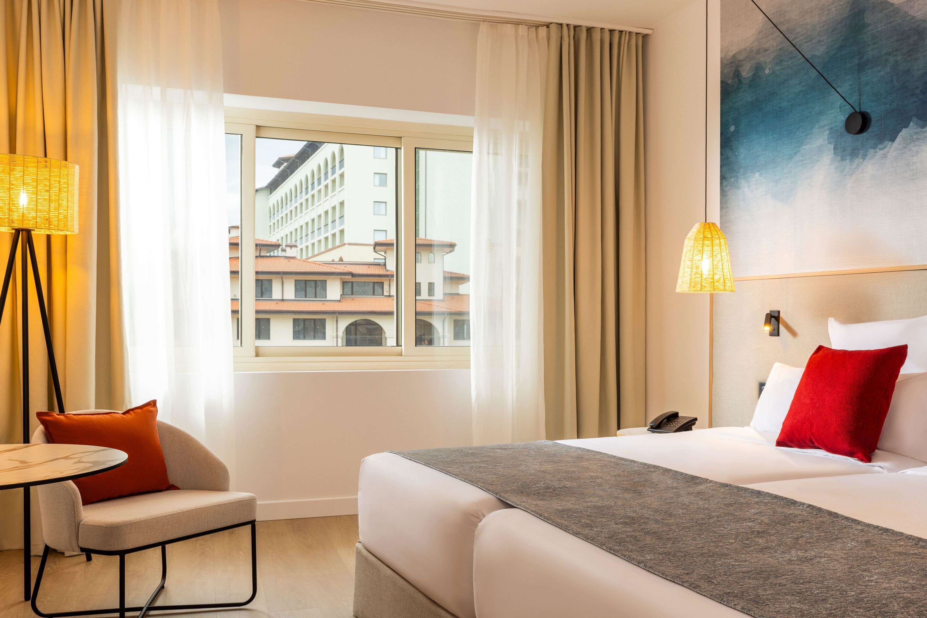 A hotel room with a bed, a chair, a table, a window with curtains, and a view of buildings outside.