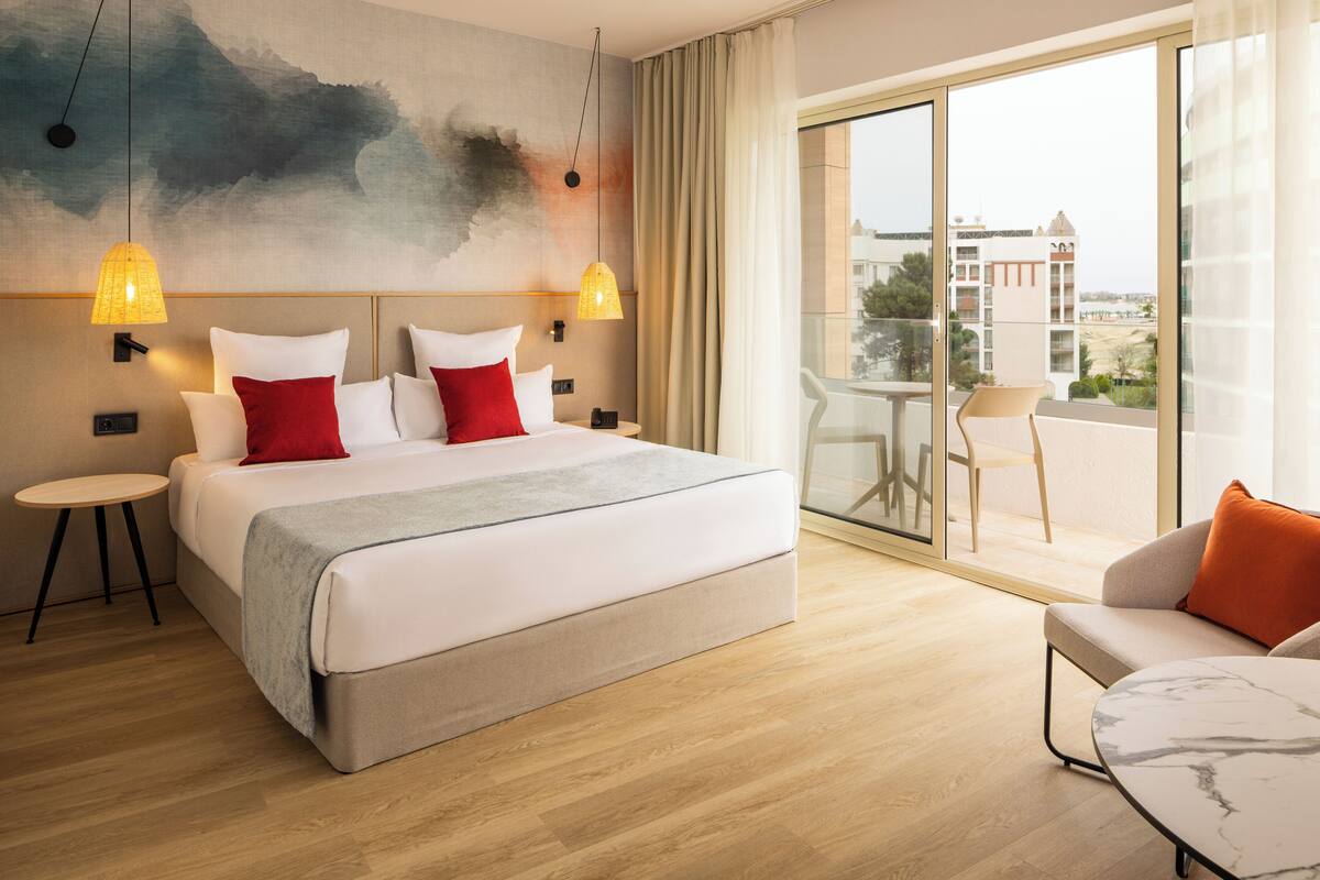 A modern hotel room with a large bed, two bedside lamps, a round table, and a balcony with a view.