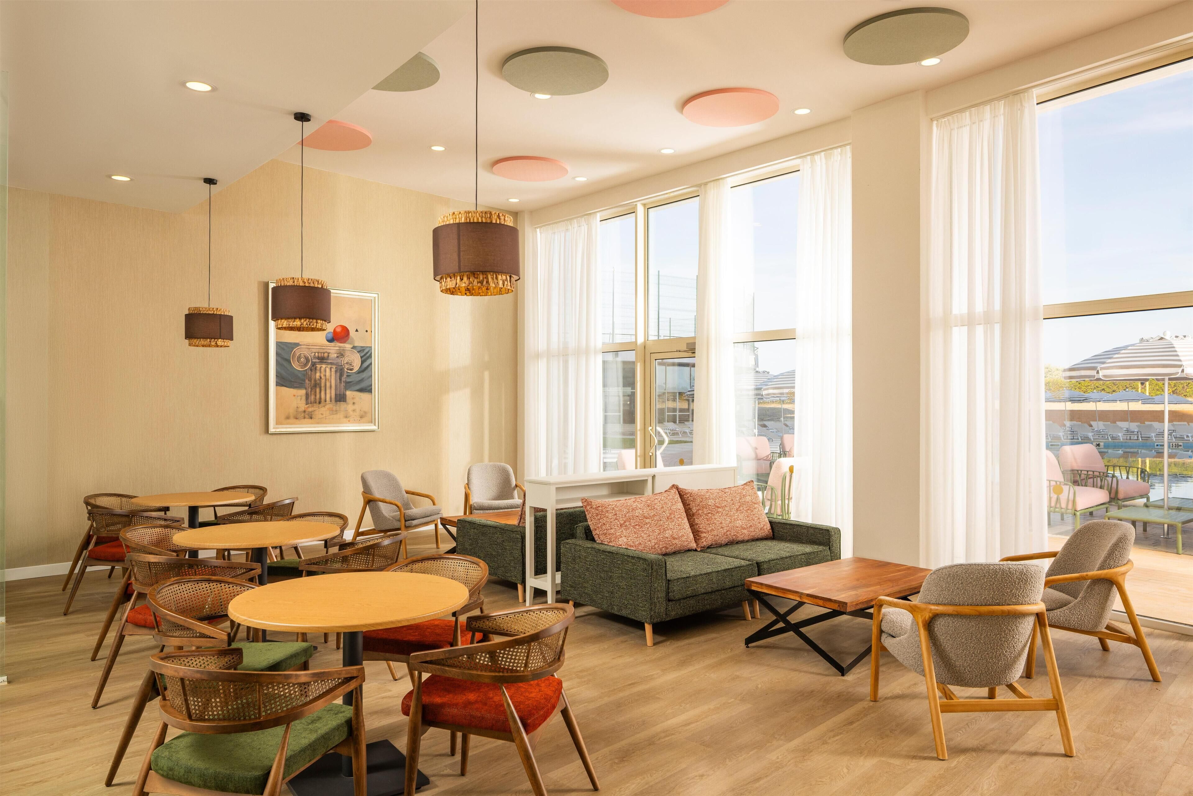 A spacious room with round tables and chairs, a sofa, and large windows offering a view of the outside.