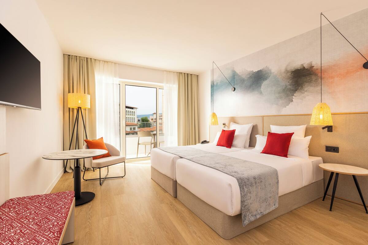 A hotel room with a bed, a desk, a chair, a TV, and a view of the outside.