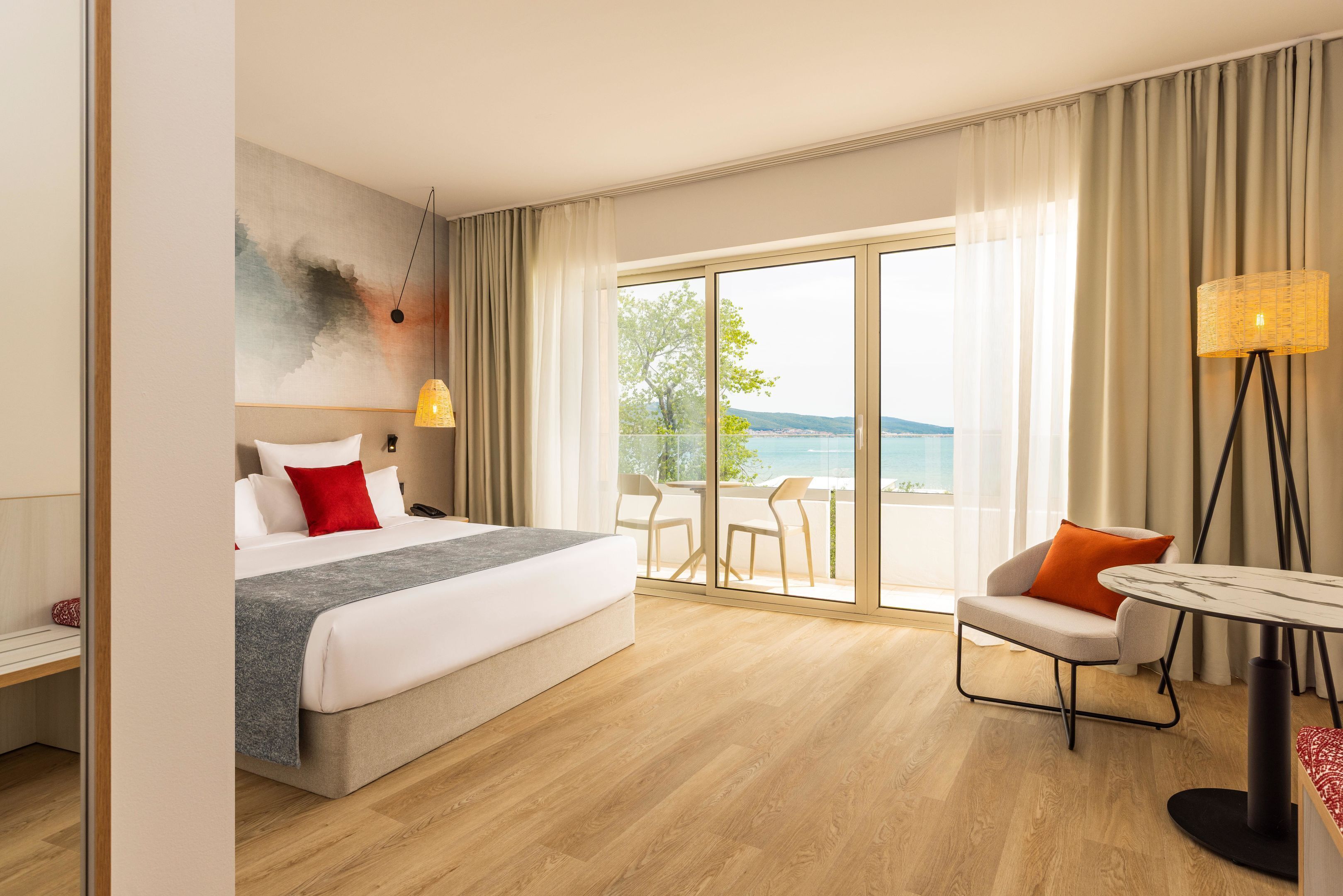 A hotel room with a bed, a desk, a chair, a table, and a view of the sea.