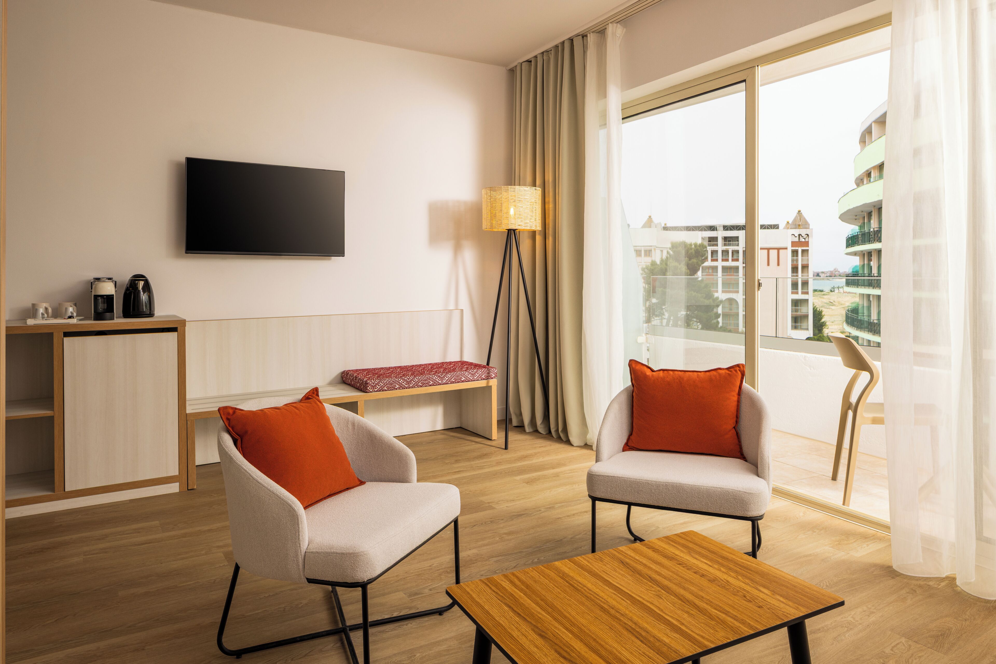Et moderne hotelværelse med et fladskærms-TV, to lænestole med orange puder, et træbord, en gulvlampe og et stort vindue med udsigt til bygninger.