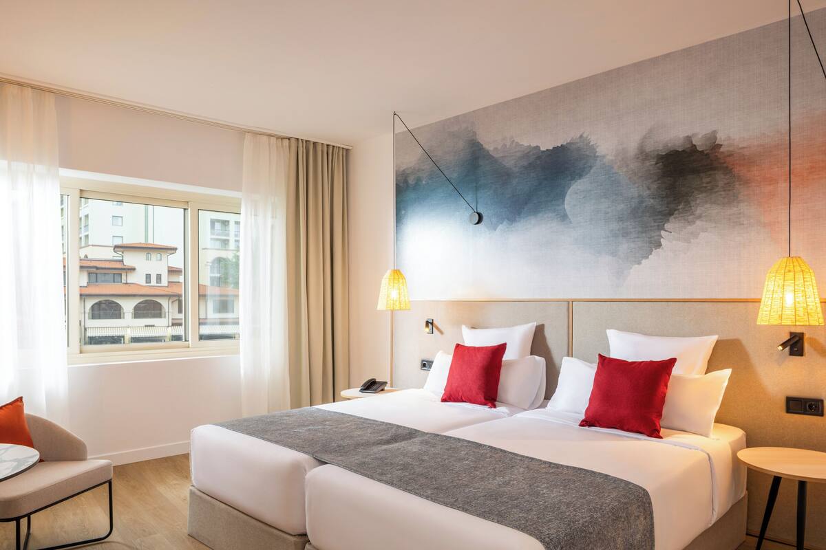 A hotel room with a large bed, two bedside tables, a chair, a window with curtains, and a wall painting.