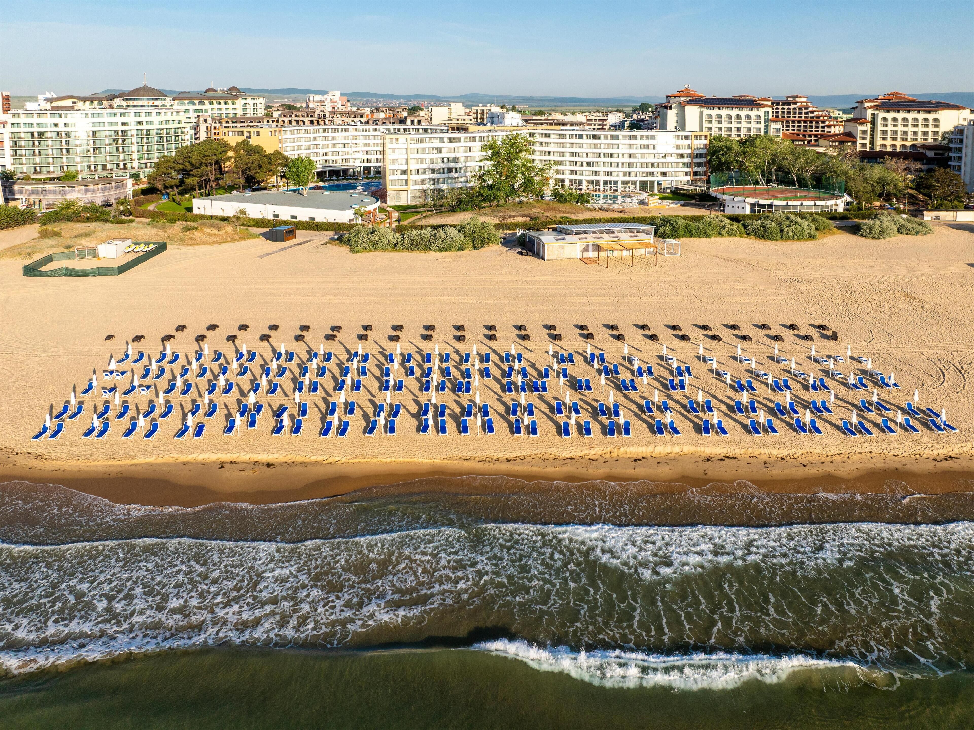 A beach with rows of blue lounge chairs and umbrellas, a building complex in the background, and waves approaching the shore.