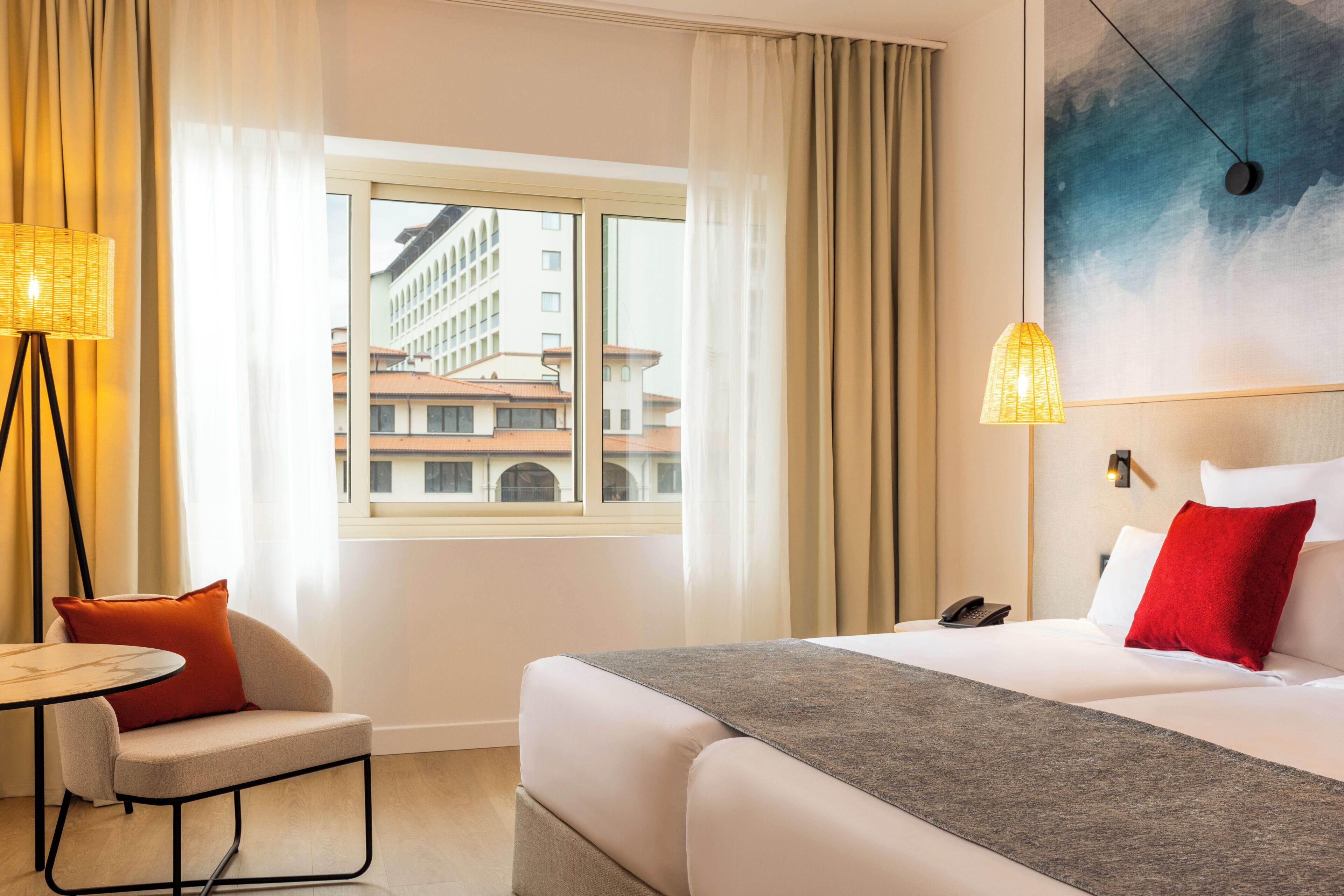 A hotel room with a bed, a chair, a table, a phone, and a view of buildings through a window.