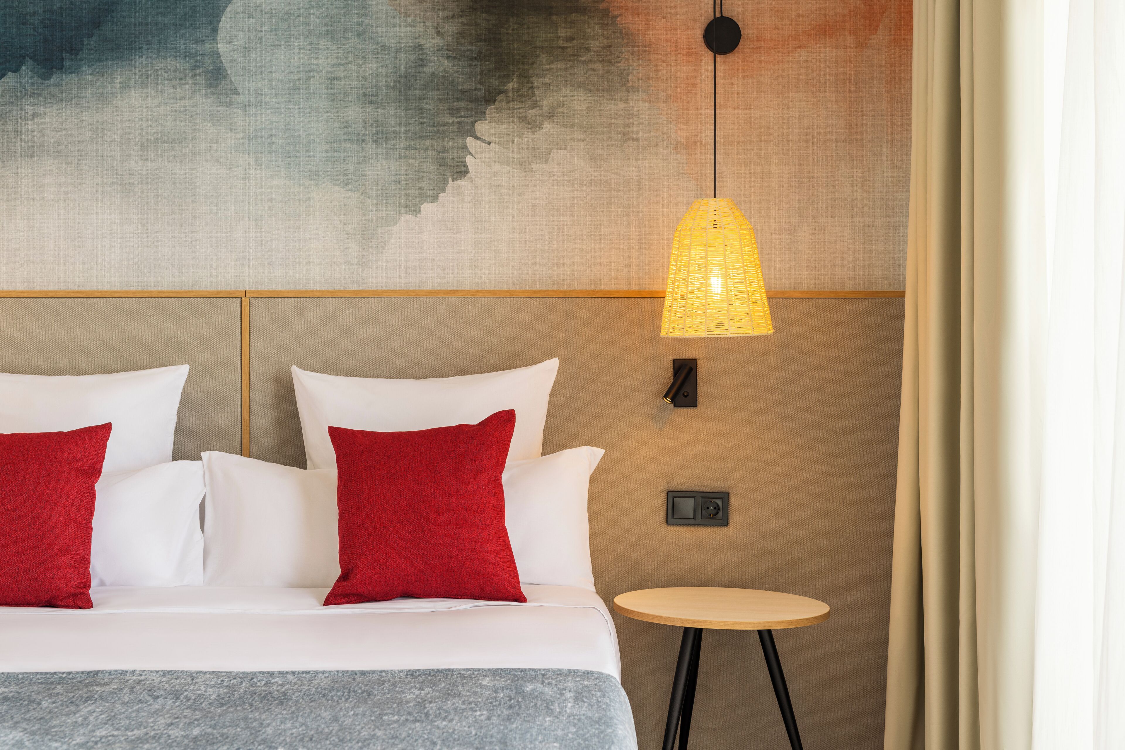 A hotel room with a bed, red and white pillows, a small round table, a wall-mounted lamp, and a textured wall.