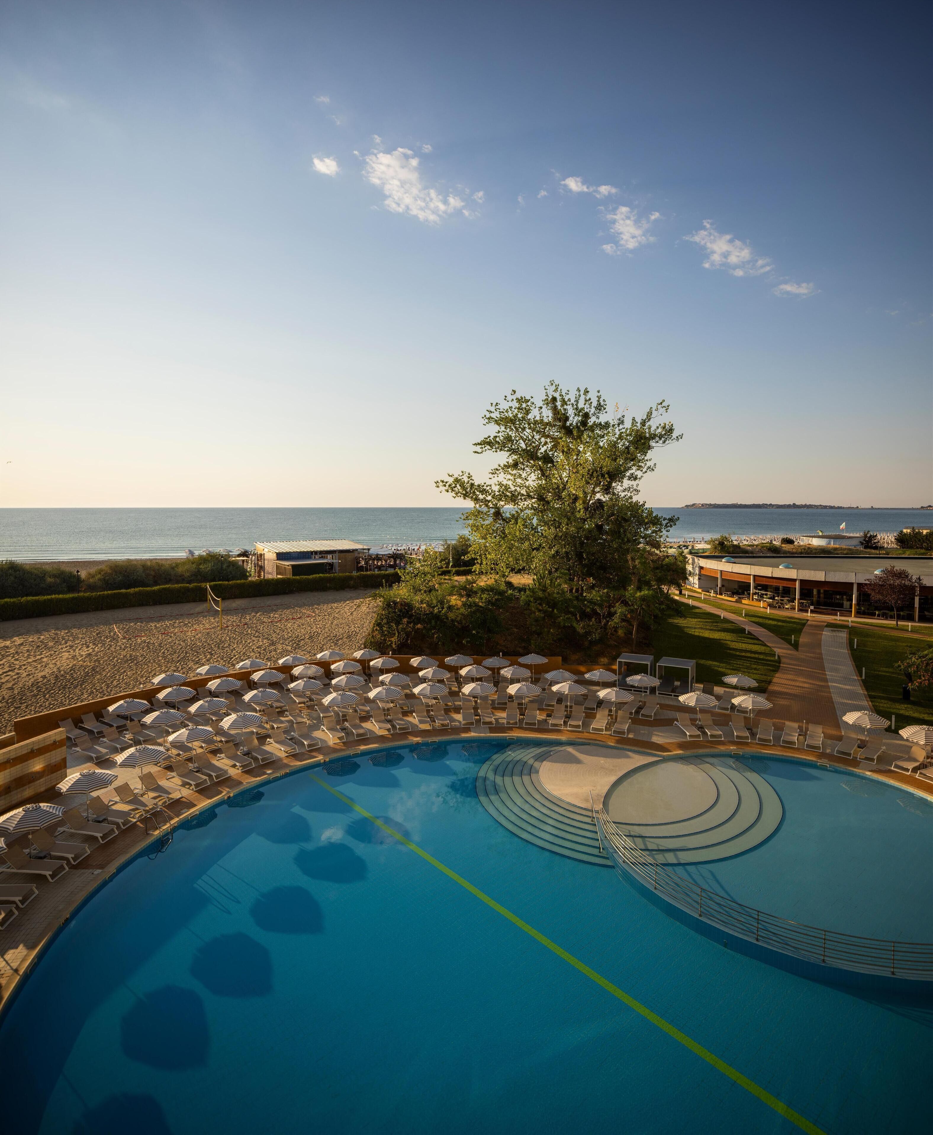 Ein Poolbereich mit Liegestühlen und Sonnenschirmen, mit Blick auf das Meer und einem angelegten Garten.