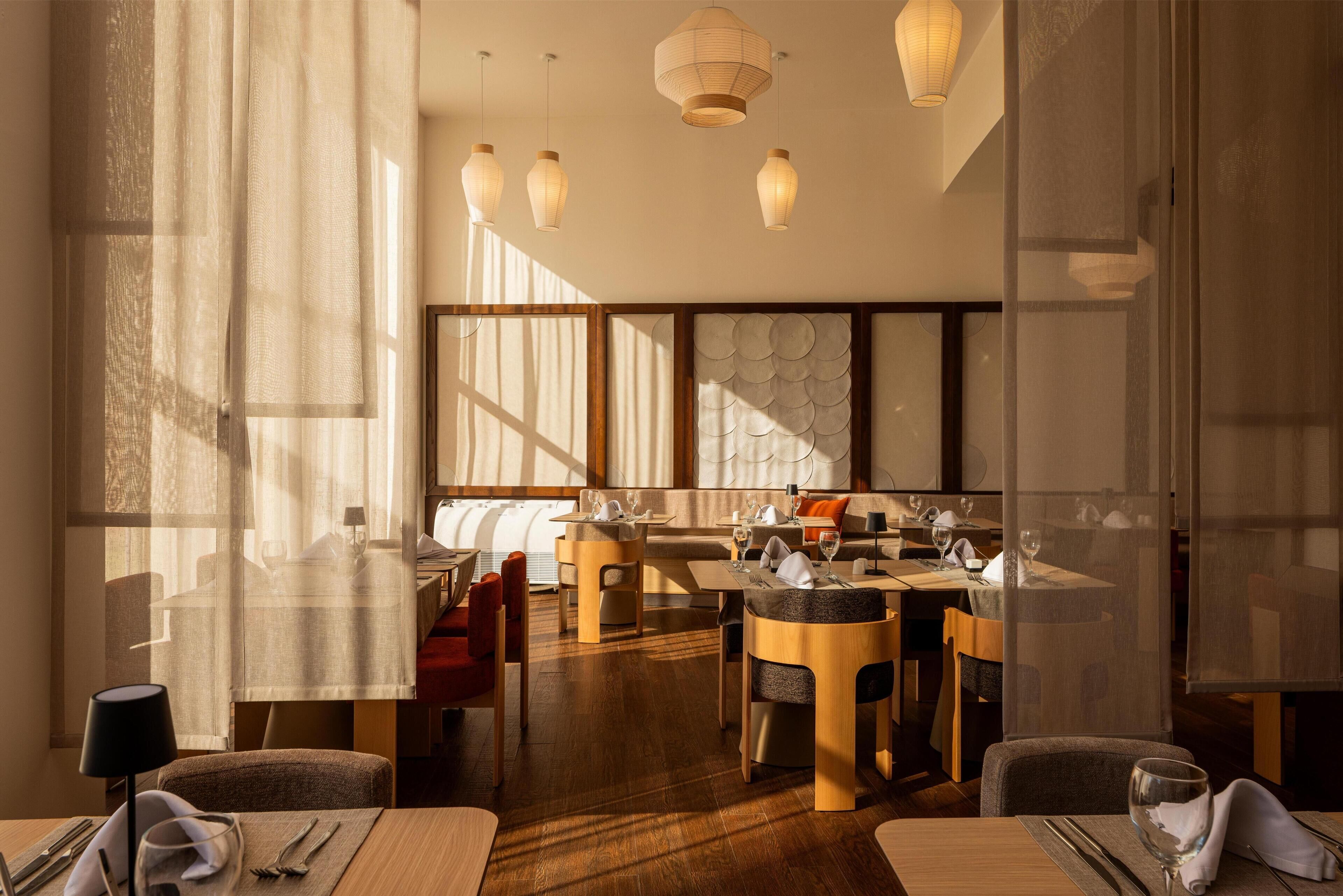 A modern dining area with wooden tables and chairs, large windows allowing natural light, and hanging pendant lights.