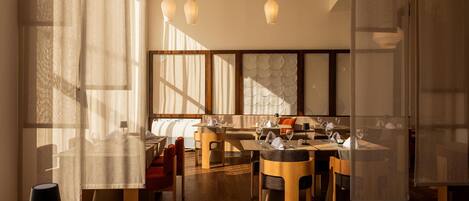 A modern dining area with wooden tables and chairs, large windows allowing natural light, and hanging pendant lights.