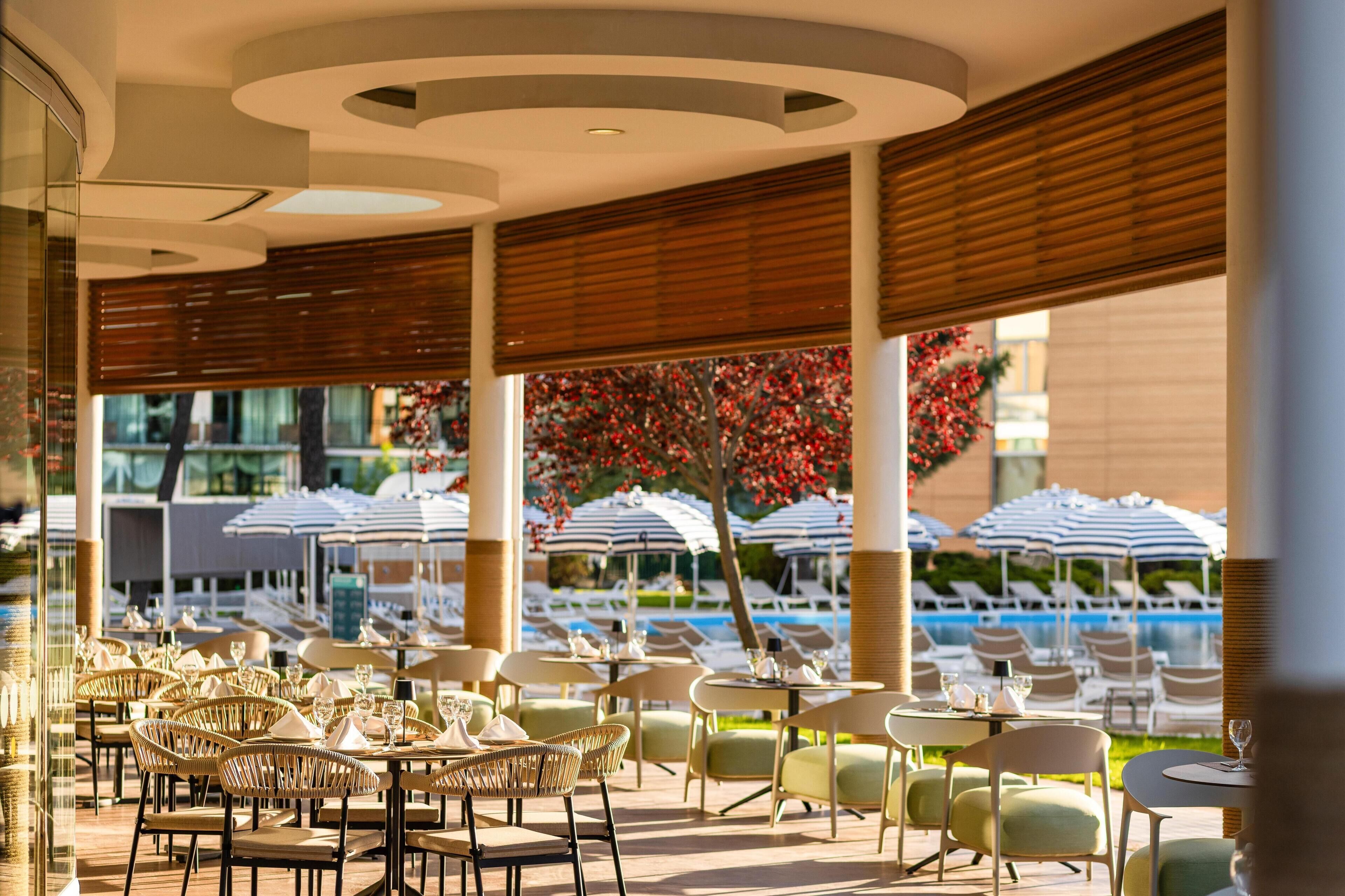 Un área de comedor al aire libre con mesas y sillas, sombrillas y una piscina al fondo.