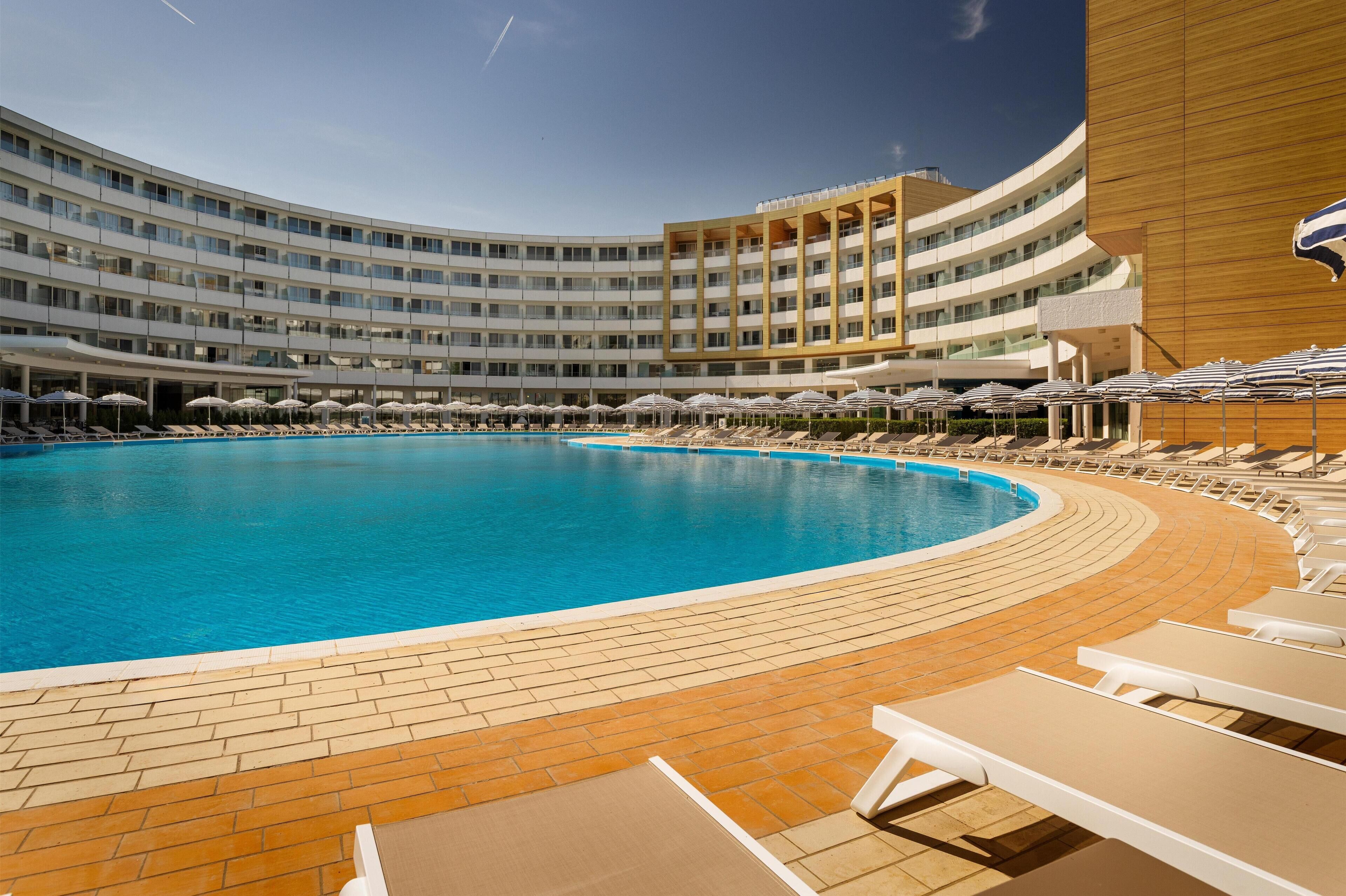 A large, curved swimming pool with lounge chairs and umbrellas, surrounded by a multi-story hotel building.